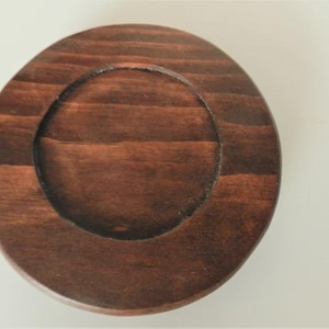 May include: A round, wooden coaster with a dark brown stain. The coaster has a recessed center, creating a lip around the edge. The wood grain is visible, adding texture to the coaster.