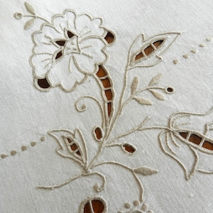 May include: White linen tablecloth with delicate embroidered floral design. The embroidery features a large flower with intricate cutouts and smaller flowers and leaves.