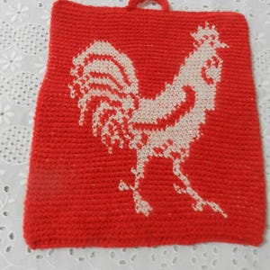 May include: A red, square, knitted hot pad featuring a white rooster design. The hot pad has a small loop for hanging. The rooster is depicted in a detailed, stylized manner, with a focus on its feathers and form.