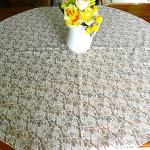 May include: A round, cream-colored lace tablecloth with a floral pattern. A white ceramic pitcher holds a bouquet of yellow and orange roses. The tablecloth is draped over a wooden table.