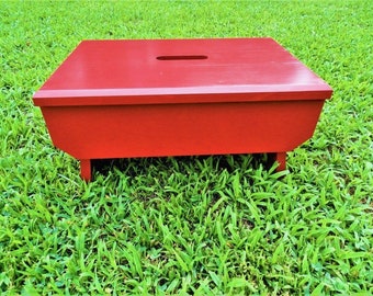 May include: A red wooden step stool with a rectangular top and a slot cut into the top surface. The stool has four short legs and is painted a solid, vibrant red. It is sitting on a bed of green grass.