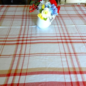 May include: A white tablecloth with a red and beige plaid pattern. The tablecloth is laid out on a table with a white pitcher and a bouquet of flowers in the center.