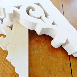 May include: Unfinished wooden corbel with ornate, scroll-like carvings. The corbel is light in color and appears to be made of softwood. The design features a combination of curved and straight lines, creating a decorative architectural element.