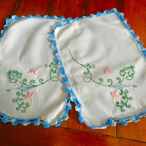 May include: Two white embroidered placemats with a blue crocheted border. Each placemat features a floral design with pink and green embroidery. The placemats are rectangular with scalloped edges.
