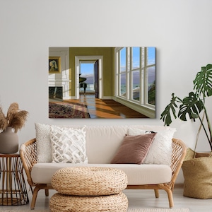 Op de afbeelding: Een ingelijst schilderij van een kamer met uitzicht op de oceaan. De kamer heeft een open haard, een deuropening naar een balkon en een groot raam. Een beige bank met kussens en een geweven poef staan op de voorgrond.