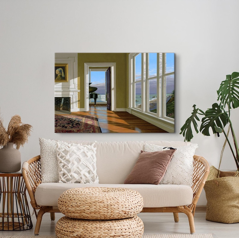Op de afbeelding: Een ingelijst schilderij van een kamer met uitzicht op de oceaan. De kamer heeft een open haard, een deuropening naar een balkon en een groot raam. Een beige bank met kussens en een geweven poef staan op de voorgrond.