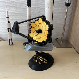 Könnte beinhalten: Ein 3D-gedrucktes Modell des James-Webb-Weltraumteleskops mit einem goldfarbenen sechseckigen Spiegelsystem und einem schwarzen Sockel mit dem Text "James Webb Space Telescope 12/25/2021".