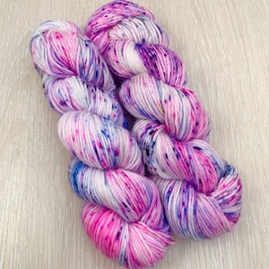 Può includere: Due gomitoli di filato tinto a mano su una base bianca con macchie rosa, viola e blu.
