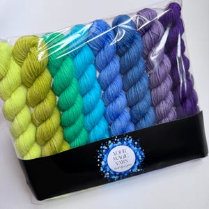 May include: A set of seven hand-dyed yarn skeins arranged in a gradient from lime green to deep purple, packaged in a clear plastic bag with a black label featuring the text YOUR MAGIC YARN hand dyed yarn in a blue circular design.