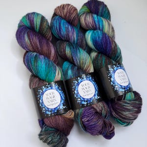 May include: Three skeins of hand-dyed yarn in shades of teal, purple, and brown. Each skein has a black label with the text "YOUR MAGIC YARN hand dyed yarn" in white. The yarn is arranged on a white surface.