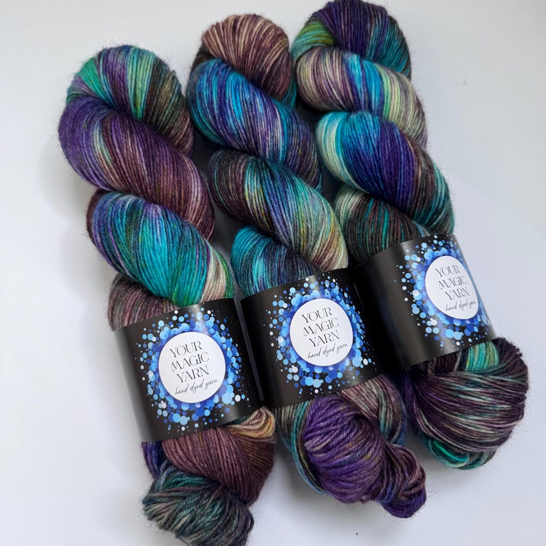 May include: Three skeins of hand-dyed yarn in shades of teal, purple, and brown. Each skein has a black label with the text "YOUR MAGIC YARN hand dyed yarn" in white. The yarn is arranged on a white surface.