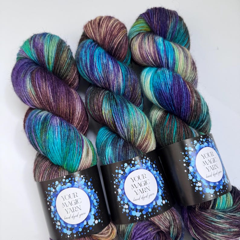 May include: Three skeins of hand-dyed yarn in shades of teal, purple, and brown. Each skein has a black label with the text "YOUR MAGIC YARN hand dyed yarn" in a white circle surrounded by blue dots. The yarn is tightly wound.