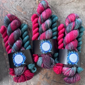 May include: Three skeins of hand-dyed yarn featuring a variegated colour palette of vibrant magenta, deep teal, and soft grey. Each skein is secured with a black paper band displaying the text Your Magic Yarn in a circular blue graphic.