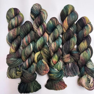 May include: A selection of hand-dyed yarn skeins. The yarn features a blend of colours, including greens, browns, yellows, and purples. The skeins are arranged in a row, displaying the variegated colour transitions. Suitable for knitting and crochet projects.