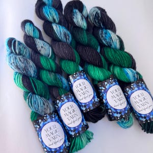 May include: Skeins of hand-dyed yarn in shades of teal, green, and black. Each skein is labeled with a circular tag that reads "YOUR MAGIC YARN hand dyed yarn". The yarn is arranged on a white surface.