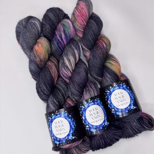 May include: Three skeins of hand-dyed yarn in shades of gray, purple, pink, and orange. Each skein has a black label with the text "YOUR MAGIC YARN hand dyed yarn" in white. The yarn is arranged on a white surface.
