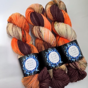 May include: Three skeins of hand-dyed yarn in shades of orange, brown, and beige. Each skein is wrapped with a black label that reads "YOUR MAGIC YARN hand dyed yarn" in white and blue. The yarn is arranged on a white surface.
