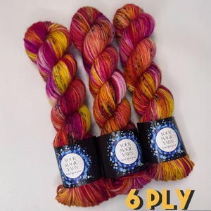 May include: Three skeins of hand-dyed yarn in vibrant colours. The yarn features a blend of yellow, orange, and magenta hues. Each skein has a black label with the text "YOUR MAGIC YARN hand dyed yarn". The text "6 PLY" is in the bottom right corner.