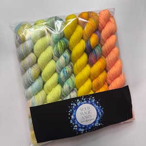 May include: A transparent bag containing several skeins of brightly coloured yarn. The yarn is arranged in rows of yellow, green, orange, and blue shades. A black label with the text "YOUR MAGIC YARN hand dyed yarn" is attached to the bag.