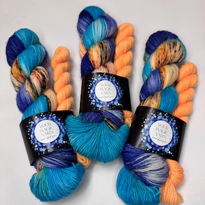 May include: Three skeins of hand-dyed yarn in vibrant colours. The yarn features a blend of blues, turquoise, orange, and yellow. Each skein is wrapped with a black label that reads "YOUR MAGIC YARN hand dyed yarn."
