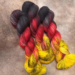 May include: Three skeins of hand-dyed yarn featuring a vibrant gradient color transition. The color palette shifts from deep charcoal black at the top, blending into a bright crimson red in the middle, and finishing with a sunny yellow at the bottom.