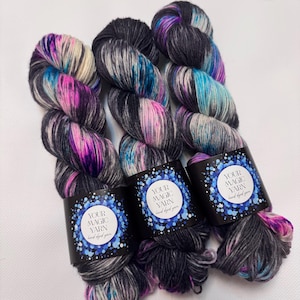 Peut inclure: Trois écheveaux de fil teints à la main, aux couleurs variées : noir, gris, rose, violet et turquoise. Chaque écheveau porte une étiquette noire avec le texte "YOUR MAGIC YARN hand dyed yarn" dans un motif bleu et blanc.