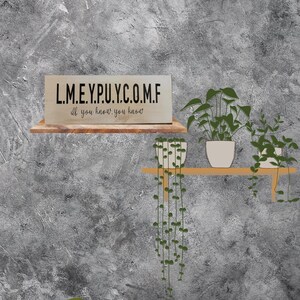 May include: A green couch with a green pillow in front of a gray wall. A wooden shelf with three potted plants is mounted on the wall. The shelf has a sign that reads "L.M.E.Y.P.U.Y.C.O.M.F. If you know, you know."