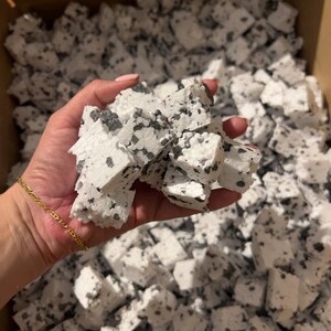 May include: A close-up of white packing peanuts with grey speckles, held in a hand. The foam peanuts are irregularly shaped. The background is filled with more packing peanuts, suggesting a large quantity of packaging material.