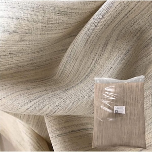 Pode incluir: Um close-up de um tecido bege com um padrão tecido sutil. O tecido está dobrado de forma que mostra a textura e o brilho sutil. Um saco de plástico transparente com um rótulo que diz "AULTICO" está no canto inferior direito da imagem.