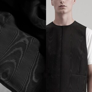 May include: Black vest with a subtle wood grain texture. The vest has a round neckline and a single pocket. The fabric appears to be a dark, solid color. The vest is worn over a white t-shirt.