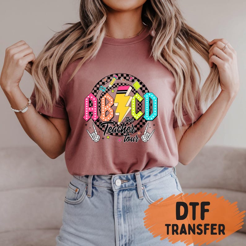 May include: A mauve t-shirt featuring a colorful graphic design. The design includes the letters ABCD in vibrant colors, a lightning bolt, and the text "the Teacher tour". The design is set against a checkered background with pencils and stars.