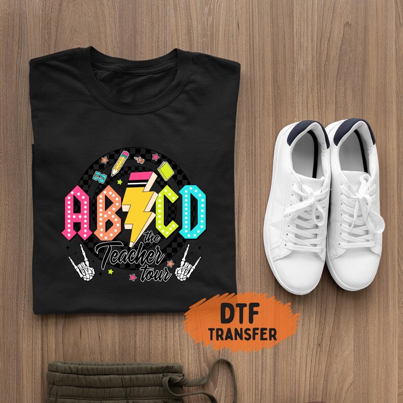 May include: Black t-shirt with a colorful graphic that reads "ABCD the Teacher Tour". The design includes a lightning bolt, pencils, stars, and skeleton hand gestures. White sneakers with navy blue accents are next to the shirt.
