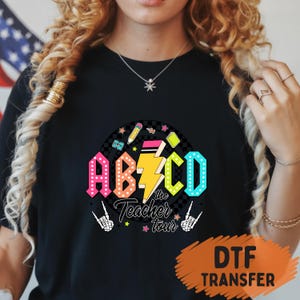May include: Black t-shirt with a colorful graphic that reads "ABCD the Teacher tour". The design includes the letters A, B, C, and D in bright colors, a yellow lightning bolt, and school-themed icons.