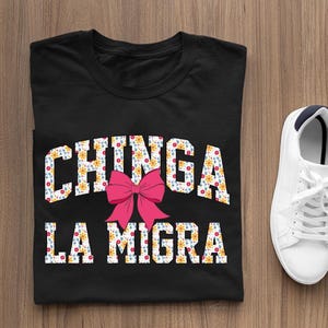 May include: Black t-shirt with a white floral print design that says "CHINGA LA MIGRA" with a pink bow in the middle.