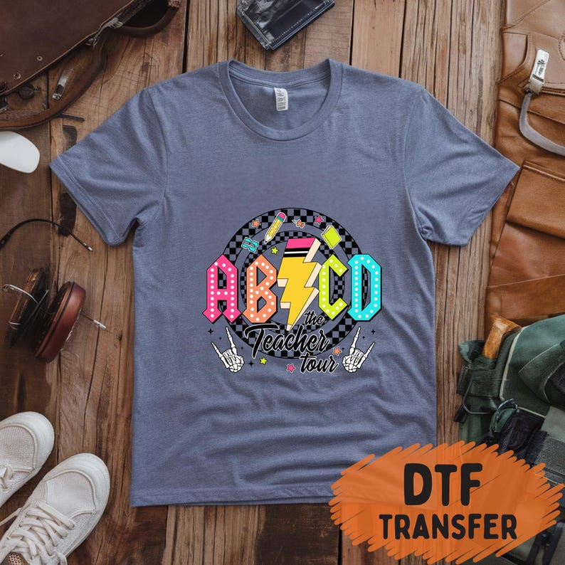 May include: A blue t-shirt with a colorful graphic that says "ABCD the Teacher Tour". The graphic includes a lightning bolt, pencils, and a checkered circle. The shirt has short sleeves and a crew neck.