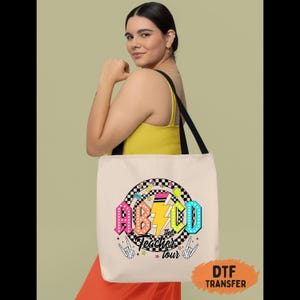 May include: A beige tote bag with black straps featuring a colorful graphic. The graphic includes the letters "ABCD" in vibrant colors, a lightning bolt, and the words "the Teacher tour". The bag is held by a person wearing a yellow top and orange pants.