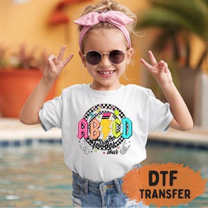 May include: A white t-shirt with a colorful graphic that says "ABCD Teacher Tour" with a lightning bolt and rock and roll hand signs. The shirt is worn by a child wearing sunglasses and a pink headband.