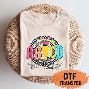 May include: A beige t-shirt with a colorful graphic design. The design features the letters ABCD in bright colors, a lightning bolt, and the words "the Teacher tour". The design includes a checkered circle and skeleton hands.