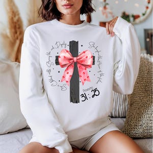 May include: White sweatshirt featuring a black cross with a pink bow. The cross has the text "She is clothed with strength and dignity" and "Proverbs 31:25" on it.