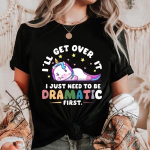 May include: Black t-shirt featuring a colourful graphic of a unicorn lying down and the text "I'll get over it I just need to be dramatic first."