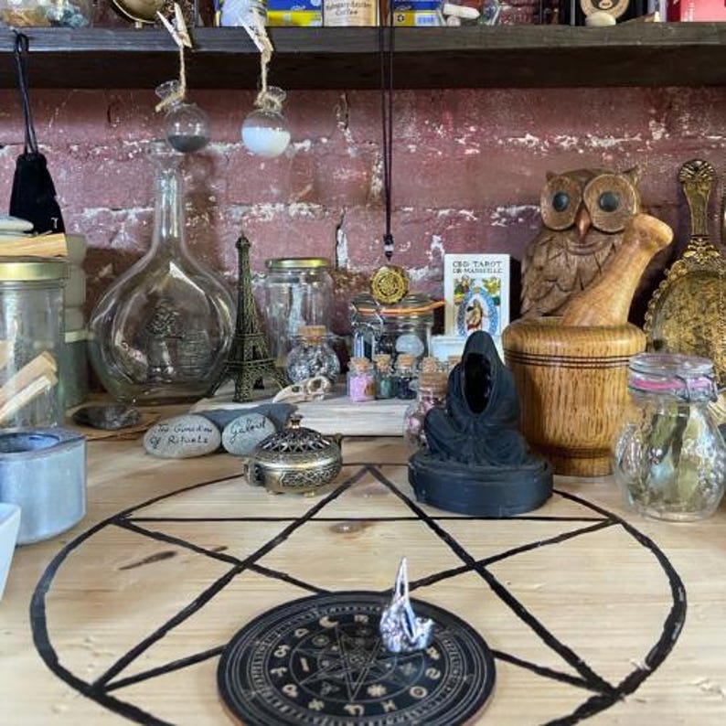 Puede incluir: Una colecci&oacute;n de objetos espirituales y ocultos, que incluye frascos de vidrio, un b&uacute;ho de madera, una estatua negra y un dise&ntilde;o de pentagrama sobre una superficie de madera. Tambi&eacute;n son visibles varias botellas peque&ntilde;as y una peque&ntilde;a Torre Eiffel.