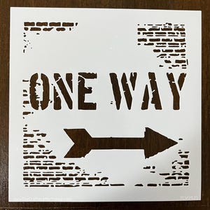 May include: A square white stencil featuring a distressed brick wall pattern in the corners and the text ONE WAY in a bold, weathered font. Below the text is a large, solid arrow pointing to the right. The stencil includes the text TCW7075.