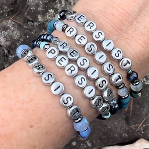 May include: Five silver bracelets with the word "PERSIST" spelled out in black letters on each bead. The bracelets are layered on a person's wrist and feature blue, green, and white beads.