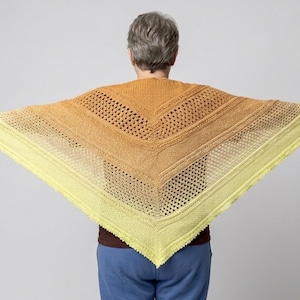 May include: A knitted shawl in a triangular shape, featuring a gradient of colors from yellow to gold. The shawl has a textured design with openwork sections and a scalloped edge. The person is wearing a brown shirt and blue pants.