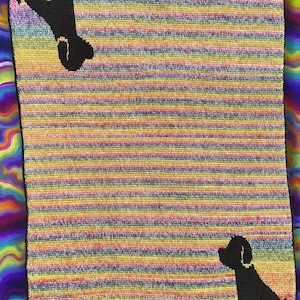 Puede incluir: Una manta a rayas multicolores con siluetas de perros negros. La manta presenta rayas horizontales en tonos de amarillo, naranja, azul y morado. Una silueta de perro está en la parte superior y otra en la inferior.