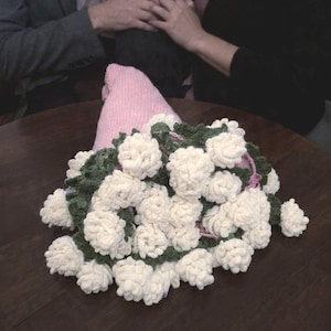 May include: A handmade bouquet of white yarn flowers with green yarn leaves and a pink knitted cone base. The bouquet is arranged on a dark brown wooden surface.