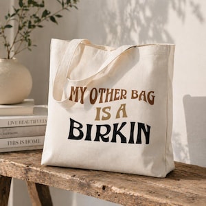 May include: Cream-colored tote bag with the text "MY OTHER BAG IS A BIRKIN" in brown and black lettering. The bag has a simple design with two handles and is made of a sturdy fabric.