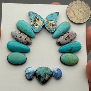 May include: A collection of eleven loose turquoise gemstones in various shapes and sizes, including a butterfly-shaped piece, arranged on a white surface next to a United States quarter coin for scale. The stones feature blue, green, and brown hues.