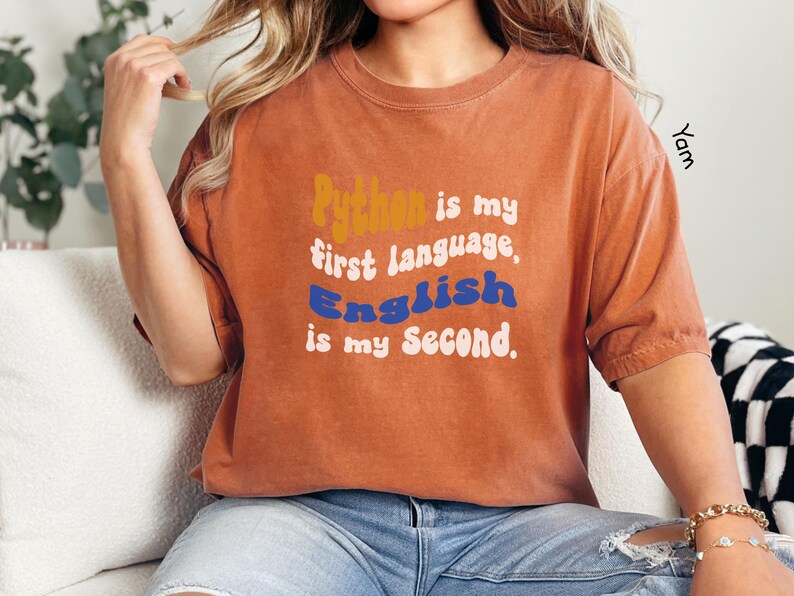 May include: A rust-colored t-shirt with the text "Python is my first language, English is my second." in a retro font with orange, blue, and white colors.