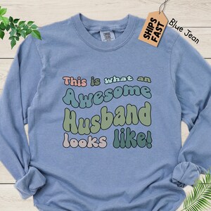 May include: A light blue long-sleeved t-shirt with the text "This is what an Awesome Husband looks like!" in colorful, retro-style lettering.